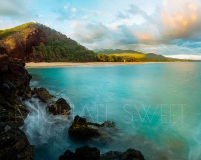 A photo taken from off shore showing a calm turquoise ocean and lush green mountains