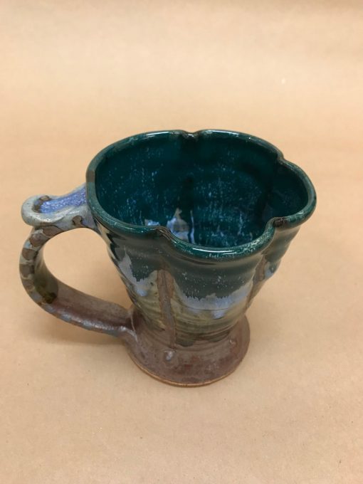 a large ceramic mug with a thumb rest and a scalloped rim. Top third of the mug is a deep green. Color changes to earthy browns with some blue in the center.