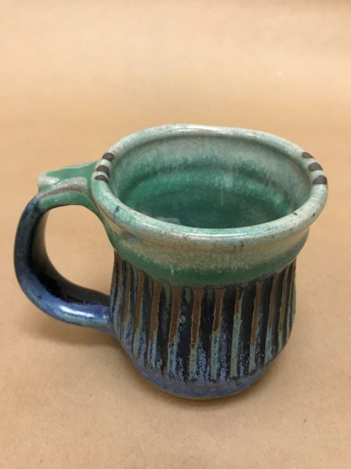 a large ceramic mug made of various blue hues, some brown colors, and aqua. Ribbed design and a thumb rest