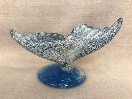 a whale tail sculpture made of blown glass. The whale tail is mostly black with white speckles, to resemble a humpback whale. On an ocean blue colored round base